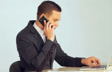 A professional man in a suit talking on a mobile phone while using a laptop to manage data for lead generation services.