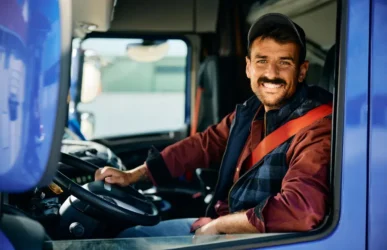 A smiling truck driver sitting in the cab of a blue semi-truck, representing the personal support and reliability provided by professional truck dispatch services.