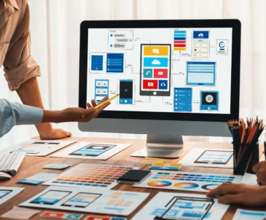 A collaborative team of designers working on a desk filled with color palettes and wireframes, pointing at a computer screen showing a mobile app UI layout as part of professional web development services.